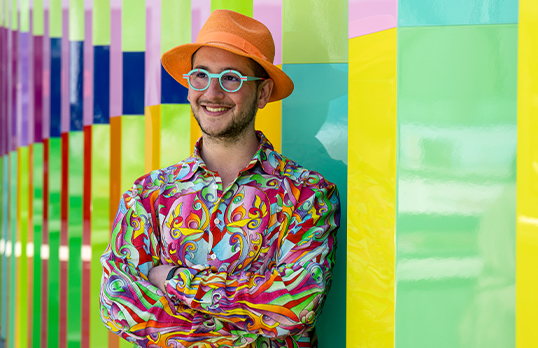 	Adam Nathaniel Furman wearing an orange hat, bright glasses and a multi-coloured shirt against an abstract background