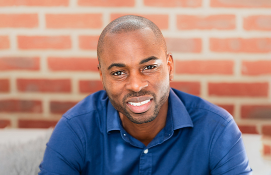 Bola Ogunmefun wearing a blue button-up shirt smiling to the camera with a brick wall background