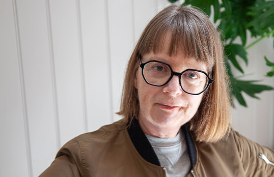 Claire Pollock wearing a brown jacket and glasses standing in front of a white panelled wall and plant