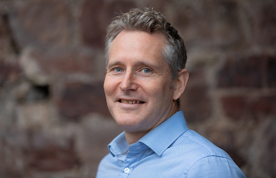 Jez Sweetland wearing a blue button-up shirt smiling to the camera with a stone wall in the background