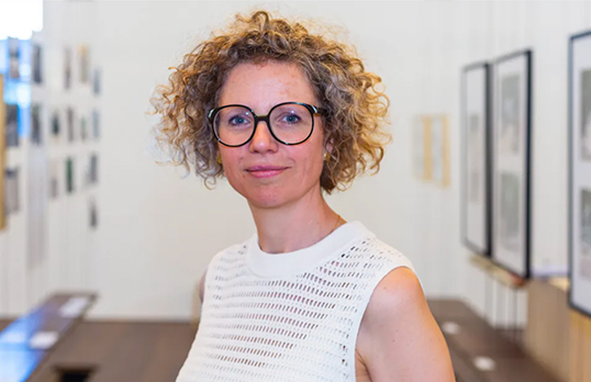 Sofie De Caigny wearing glasses and a white short-sleeve top looking at the camera with an office and hung photo frames in the background