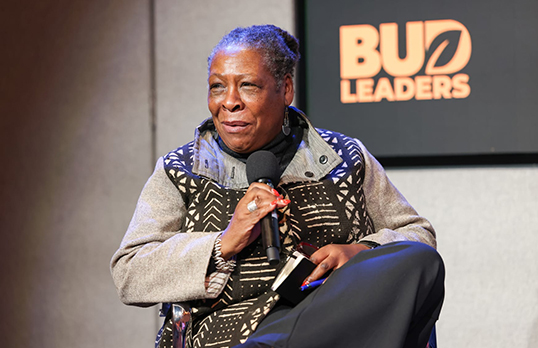 Yvonne Field portrait wearing a grey pattered coat sitting on stage holding a microphone
