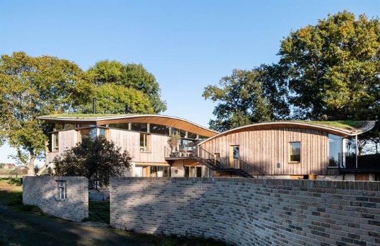 Two arched wooden clad wings of building with green roofs set amongst trees and behind a short brick wall.