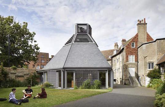Irregular shaped metal building with steep sloped roof in courtyard surrounded by old brick buildings.