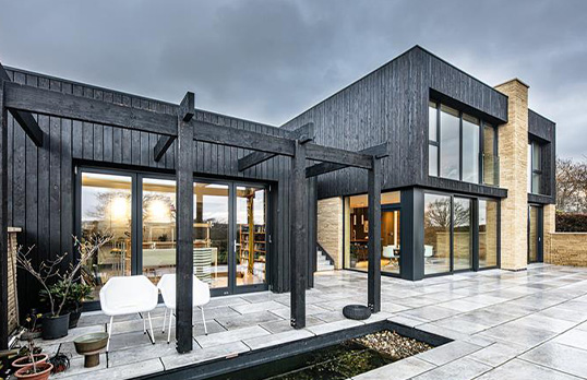 Square modern dwelling clad in vertical black timber, with large floor length windows and asymmetric height