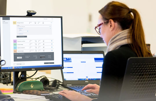 Adult with long brown hair and glasses facing a computer monitor and laptop with graphs on the screens