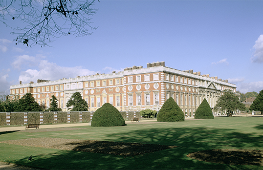 A coloured photograph of Hampton Court Palace exterior with shrubbery and grass in the foreground