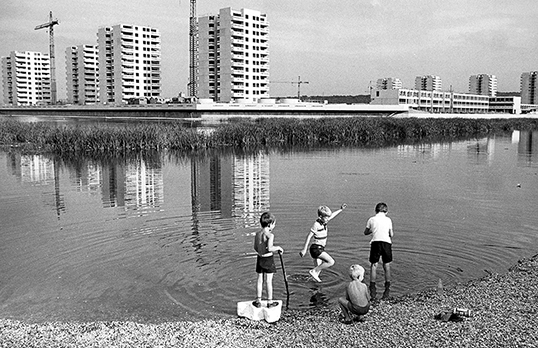 Black and white photograph of children at the edge of a shallow lake with blocks of flats and cranes in the background 