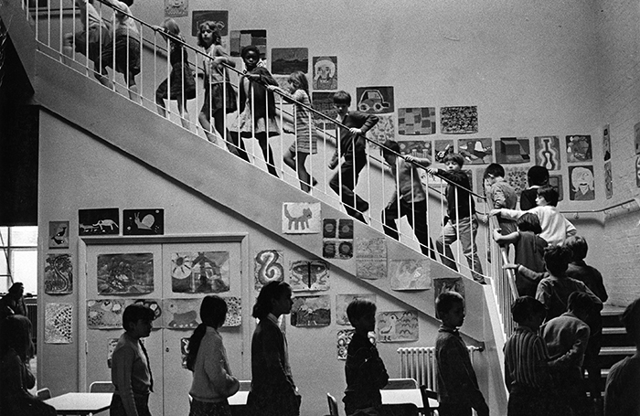 Black and white photograph of school children in a queue walking up a set of stairs with photos on the wall