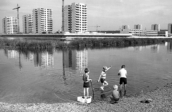 Black and white photograph of children at the edge of a shallow lake with tower blocks and cranes in the background