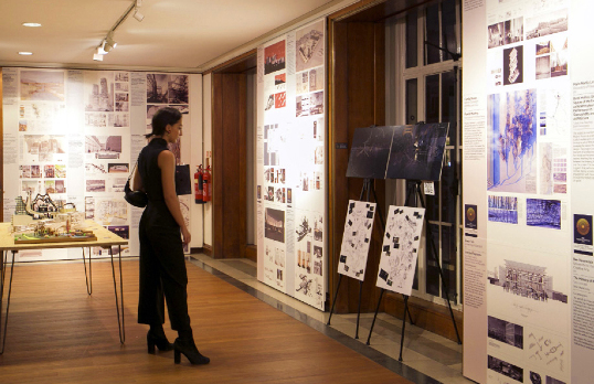 Adult woman wearing all black, looking at an exhibition panel made up of text and images covering the walls and stand board in front of a window