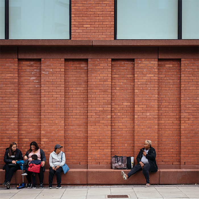 Parents sitting on a low bench in front of a brick wall waiting to pick their children up from school - Kingsland