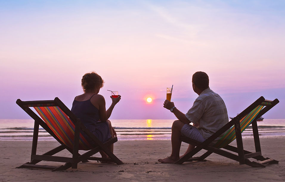 a man and woman sitting on deckchairs at the edge of the ocean watching the sun set over a purple sky