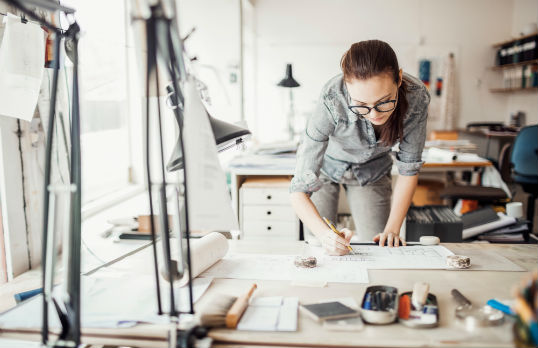 Adult with long hair and glasses leaning over a architectural drawing in a office setting