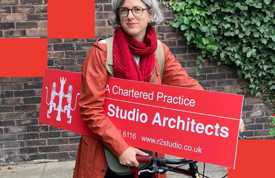 Lady with orange coat and bike holding red RIBA signboard