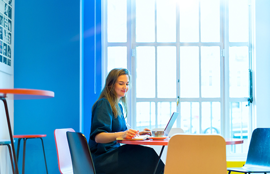 Worker with long hair using laptop at a table next to a window in a blue room