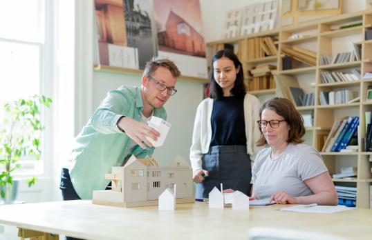Three adults sitting and standing around a table, one adult is holding part of a white housing model 