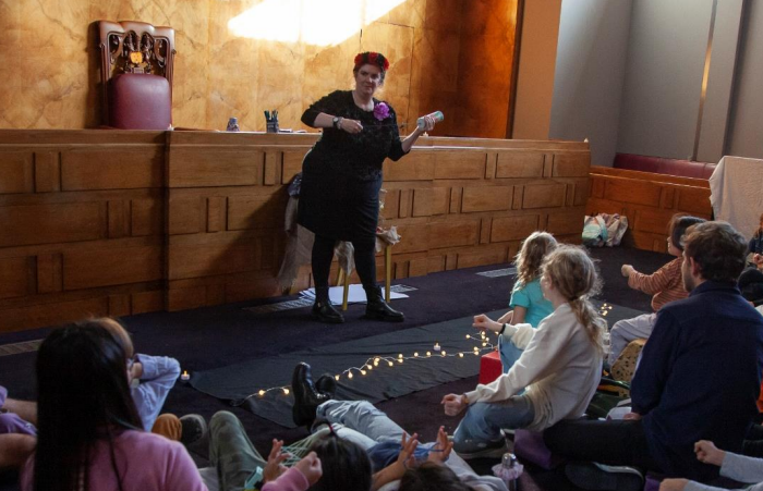 A woman in a black outfit and flower crown pretending to play the guitar in front of a group of schoolchildren