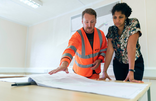 A man in a high vis jacket and a woman looking at blueprints
