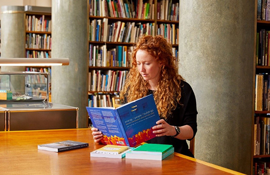 Woman with ginger hair sitting in a library reading a book