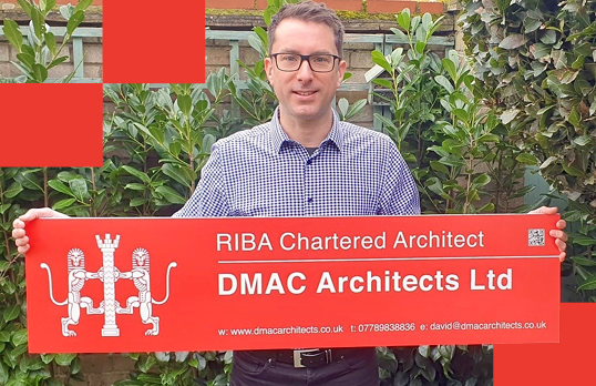 Man with glasses holding RIBA Signboard with greenery behind