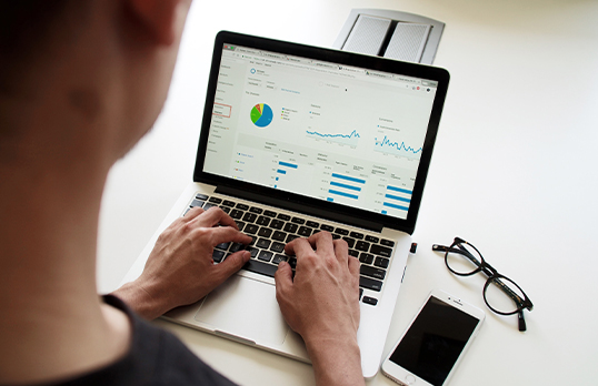 View over an adults shoulder who is typing on a laptop with graphs on the screen, and a pair of glasses and iPhone next to the laptop on the table