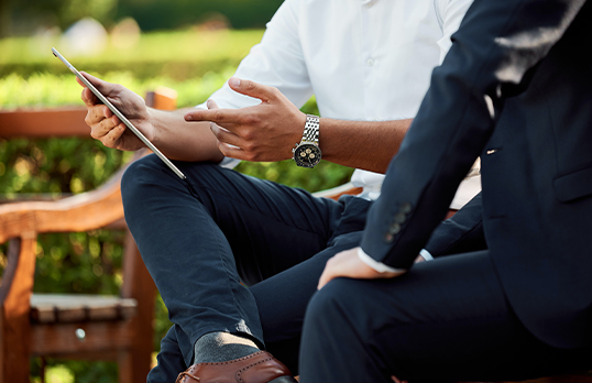 Two adults in suits sitting on park benches facing each other, with one crossing their legs and pointing to a tablet