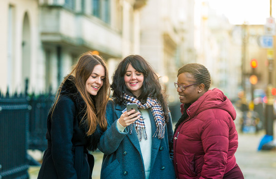 Three adults standing on a street in winter coats looking a cellphone and smiling
