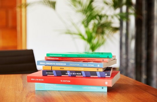 Stack of brightly colour RIBA books on a wooden table with plant in the background