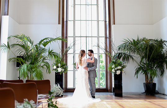 Couple in a wedding dress and suit embracing in front of a tall Art Deco window surrounded by palm plants