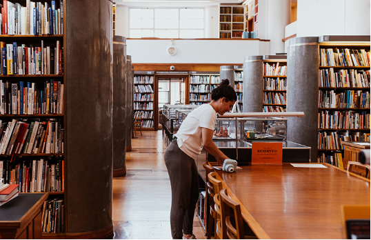 A woman with dark hair up in a bun leaning over books on a table in the middle of the RIBA Library with rows of bookshelves in the background