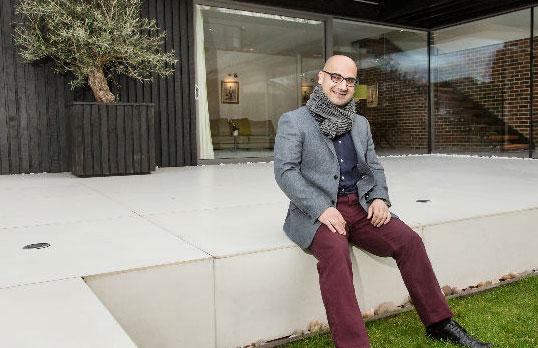Dan Benham sitting on the edge of a patio of a contemporary house made of glass and dark stained wood