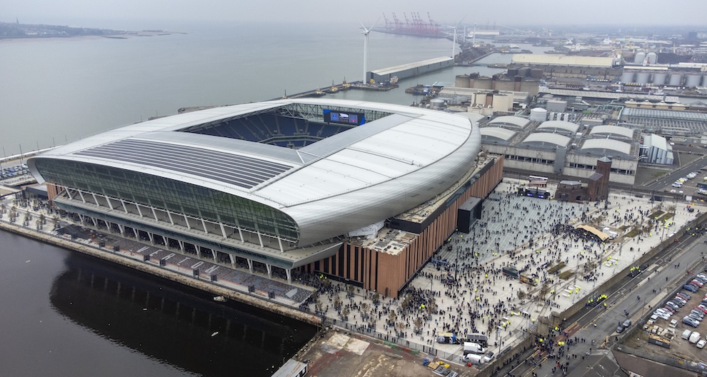 Aerial view of Everton FC stadium - a large rectangular sports ground with brown brick lower and aluminium clad stands and roof eith opening over pitch. Located beside the river with large public realm square between stadium and road