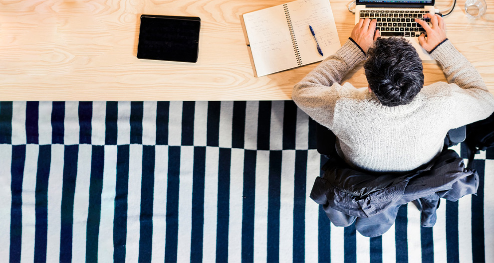 Aerial view of adult with short black hair sitting at a wooden desk with open laptop and notebooks, a blue and white striped carpet below