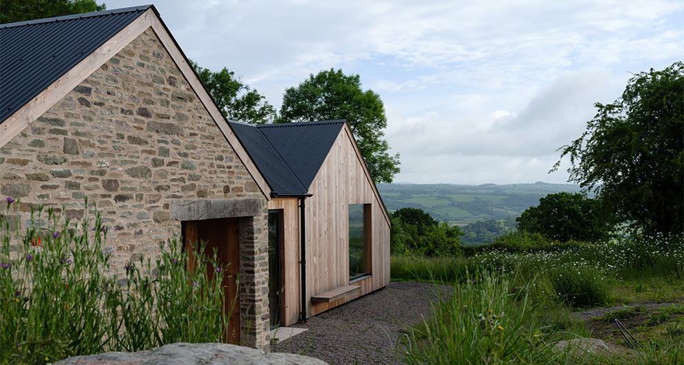 Pen Y Common stone and wood cottage overlooking grass fields and trees by Nidus Architects and Rural Office